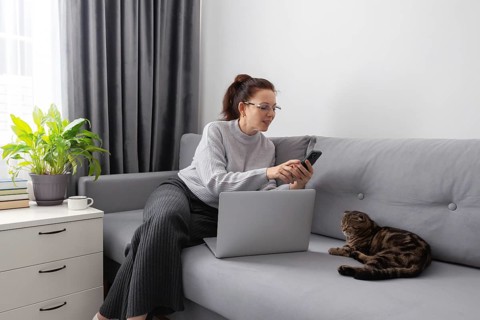 A woman works from home on a grey couch, using her laptop and smartphone. A cat relaxes beside her,