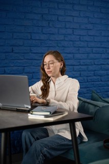 A woman in a white shirt and glasses focuses intently on her laptop in a stylish cafe with a blue