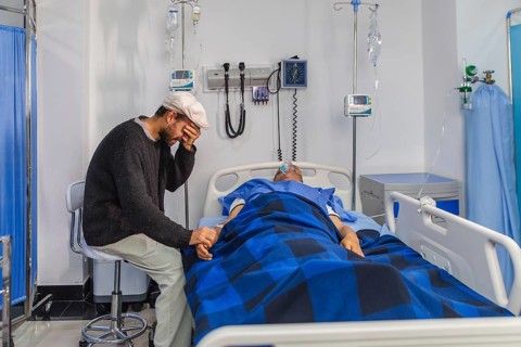 A man sits beside a hospital bed, holding the patient's hand in an emotional moment. Medical