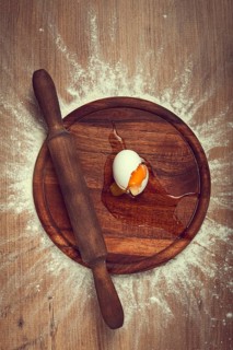 A cracked egg sits on a wooden board with a rolling pin beside it, flour scattered around,