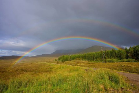 A rainbow stretches across a green meadow under a cloudy sky in a rural landscape, Lairg, Scotland,