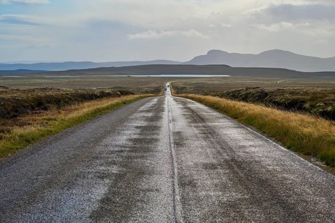 Deserted road leads through vast landscape with mountains in the background and overcast sky,