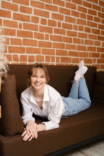 A young woman lies on a brown sofa, smiling. She wears a white shirt and light blue jeans. The