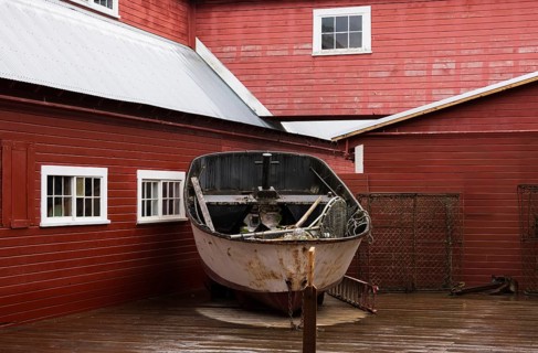 An old, weathered boat stands on display against a vibrant red wooden building in Hoonah, Alaska,