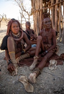 Young and old Himba woman, traditionally dressed, sit in front of a hut in the shade, near Opuwo,