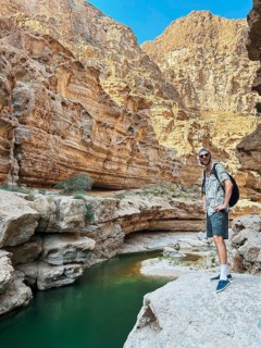 A male hiker stands on a rocky outcrop overlooking the tranquil oasis green waters of Wadi Ash Shab
