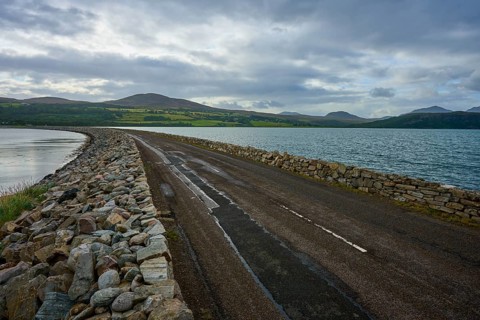 A ring road along a lake under a slightly cloudy sky with mountains in the background, Tongue