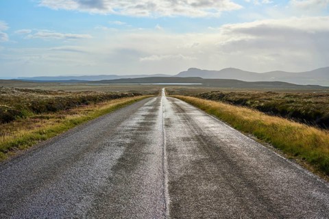 A long, empty road leads through a vast landscape of distant mountains under a cloudy sky, Tongue,