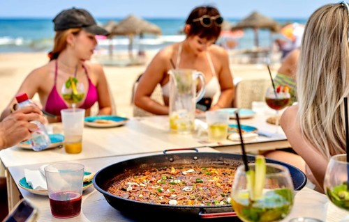 Friends gather around a table on the beach, savoring a large serving of paella surrounded by