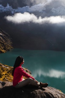 An Ecuadorian woman in a red jacket sits by a lake, enjoying the breathtaking view of El Altar
