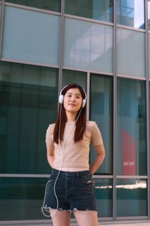 A young woman stands confidently outside a modern building, listening to music on white headphones.