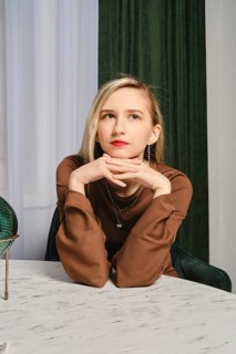 A young woman with blonde hair and red lipstick rests her chin on her hands while sitting at a