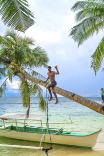 Young man making shaka sign while sitting on a palm tree swing over a tropical beach in kangbangyo