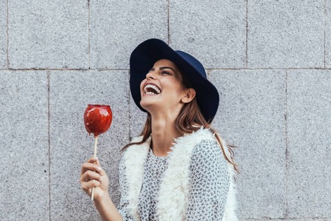 A happy woman in a stylish hat and furry vest laughs joyfully while holding a candy apple. The grey