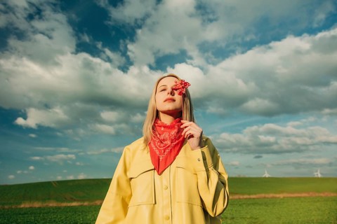 Woman in a yellow coat holding a red pinwheel, standing in a green field with a vibrant blue sky