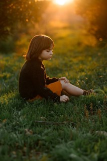 A child sits serenely in a grassy field with orange trees during sunset. The golden light creates a
