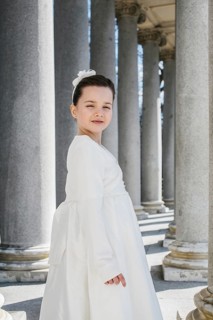 A young girl elegantly dressed in a First Communion Day dress stands in front of classic stone