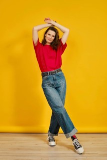 An adult woman stands confidently with her arms raised, wearing a red t-shirt and blue jeans