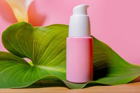 A pink skincare product bottle with a pump dispenser rests elegantly on a green, lush leaf, against