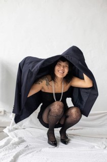 A cheerful Asian woman with meaningful tattoos crouches under a black jacket, laughing joyfully.