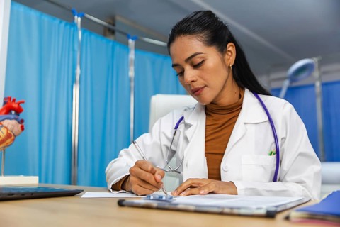 Female doctor with a stethoscope around her neck attentively writes notes at her desk in a medical
