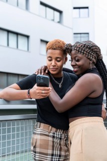 A joyful LGBT multiethnic couple embraces while looking at a phone together on a balcony, set