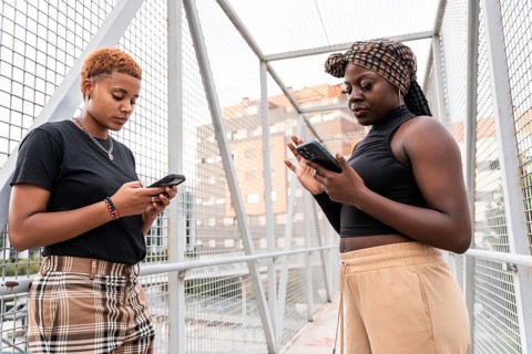 A multiethnic couple stands on a metal bridge, absorbed in their smartphones. They are casually