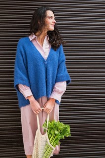 A woman in a blue cardigan and striped dress carries a net bag filled with fresh greens, standing