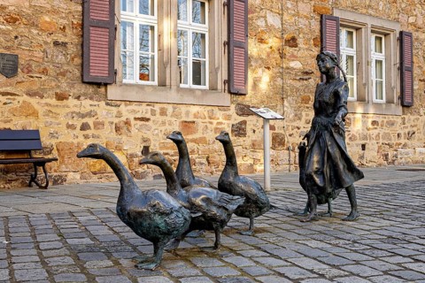 Statue of a girl with several geese in front of an old stone building with purple shutters, the