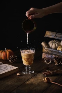 Crop anonymous person adding milk from small jug to glass with pumpkin spice latte at autumn