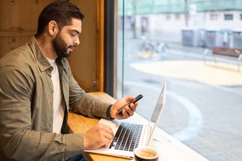 Focused individual working on a laptop while checking a smartphone in a cozy cafe setting of the