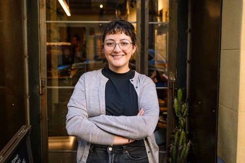 A friendly barista stands confidently at the entrance of a specialty coffee shop, exuding warmth