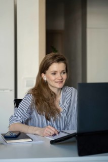 A woman sitting at her desk at home, smiling as she looks at her laptop screen. She is wearing