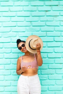 A young Latin woman in sunglasses and summer attire, smiling and holding a hat against a vibrant