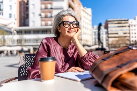 Senior woman with gray hair and glasses relaxing at an outdoor city cafe, sipping coffee and