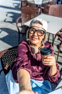 Modern woman in glasses and a cap sitting at an urban outdoor cafe, holding a takeaway coffee,