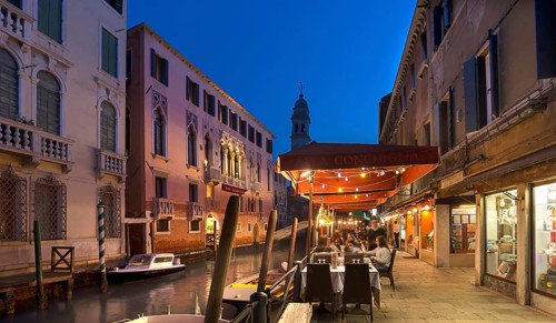 In the evening, lively restaurant on a canal with the tower of Santa Maria della Pietà in the back,