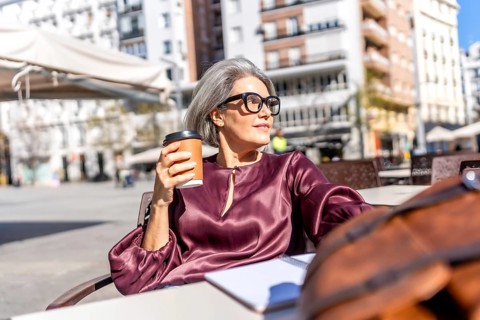 Mature woman with gray hair and eyeglasses smiling as she sits at a sunny outdoor city cafe holding