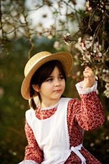 A child wearing a straw hat and vintage dress observes the blossoms on an almond tree branch. This