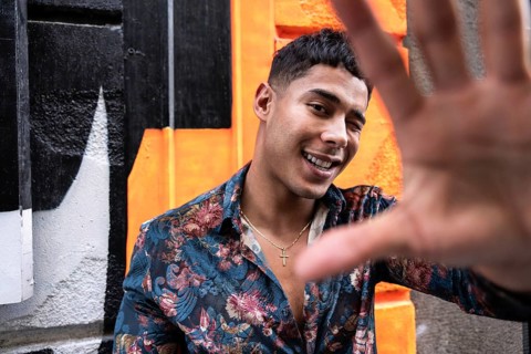 A cheerful man in a floral shirt playfully poses with his hand extended towards the camera against
