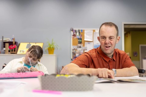 A man with a intellectual disability smiles while sitting at a table, while a woman focuses on a