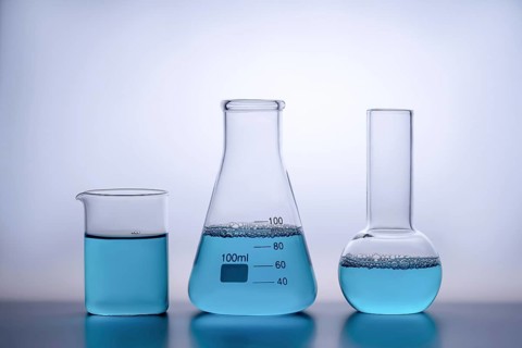 Three laboratory glassware items filled with a blue liquid sit on a reflective surface against a