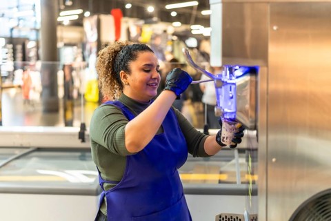 Woman in blue apron and black gloves smiles while dispensing creamy soft serve ice cream from a