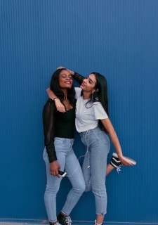 Two young friends stand against a blue wall, sharing headphones and enjoying music. They are