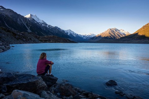 A woman is sitting on a rock at Tasman Lake. On the left, Mount Cook. Golden hour. Aoraki Mount