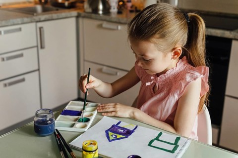 A young girl diligently paints in the kitchen using a brush and vibrant watercolors. Her artistic