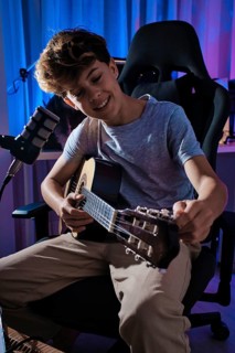 A young boy is tuning a guitar while sitting in a comfy chair, surrounded by colorful lighting. A