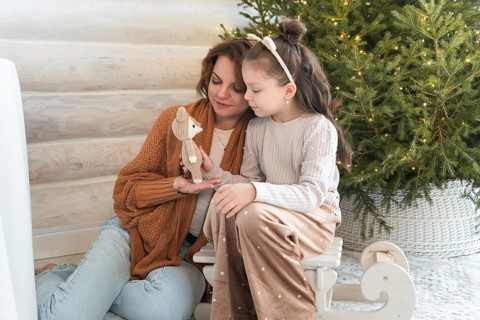 A mother and a young daughter, are sitting beside a Christmas tree, sharing a joyful moment with a