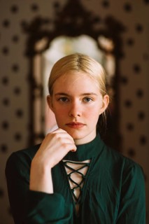 A poised young woman with blonde hair and blue eyes wears a dark green lace-up dress, gazing
