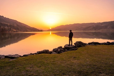 A person looks at a lake at sunset surrounded by hills and rocks, Großer Alpsee, Immenstadt im